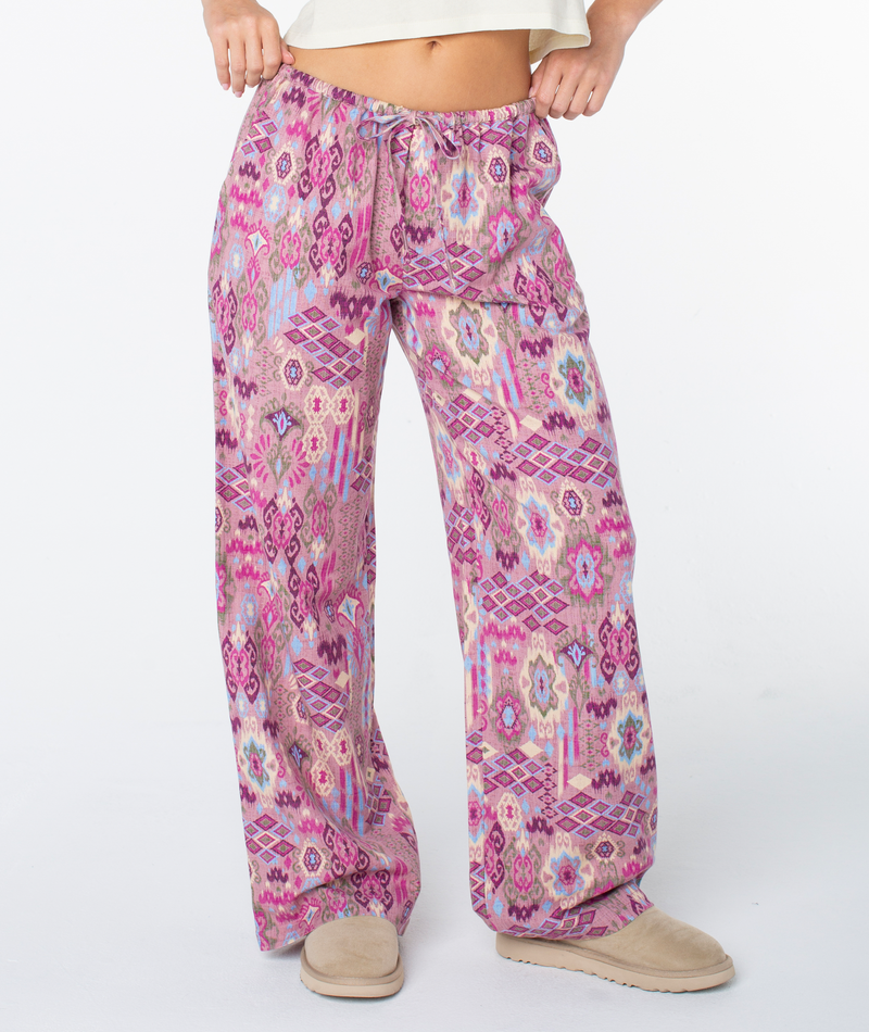 Person wearing the Roxy Iyla Pant 2, featuring a relaxed low-rise fit with colorful pink, purple, and beige patterns, paired with tan slippers. Upper body partially visible against a plain white background.