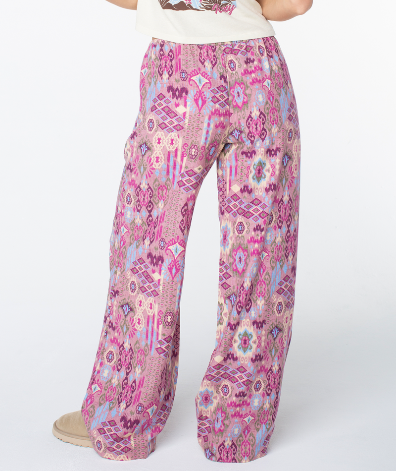 Model wearing the Roxy Iyla Pant 2—relaxed, low-rise pink pants with geometric and floral patterns—stands on a white background, showcasing their coastal-inspired comfort. Only the lower half is visible.