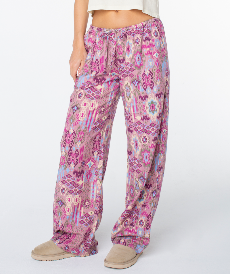 Model showcases the Iyla Pant 2 by Roxy—comfy, pink patterned pajama pants—paired with beige slippers. Only the lower body is visible, emphasizing their relaxed fit and laid-back coastal vibe.