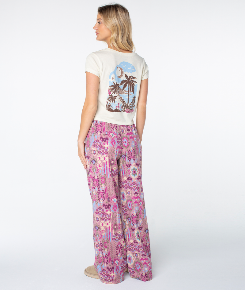 A woman faces away, wearing a cream tee with a palm tree and sun on the back and Roxy’s Iyla Pant 2 in pink and purple patterns, exuding coastal charm as she looks to her right.