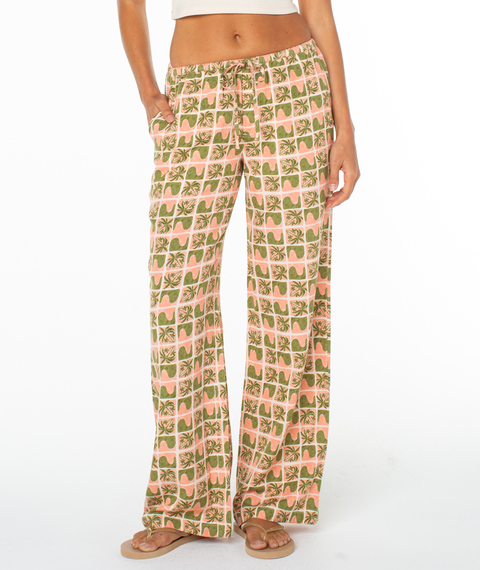 Barefoot and exuding coastal charm, a person stands with one hand in the pocket wearing the Roxy Iyla Pant 2, which features a pink and green checkered floral pattern and a relaxed low-rise fit against a white background.