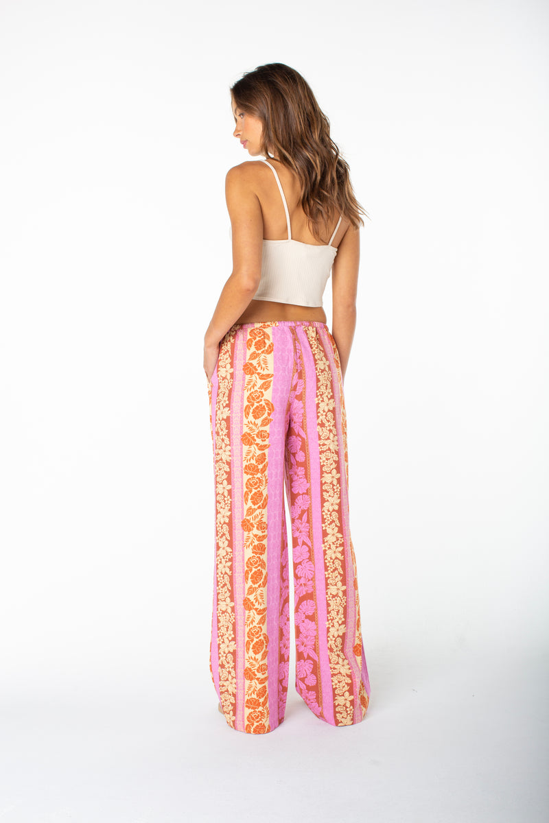 A woman stands with her back to the camera in a cream tank top and Roxy’s Iyla Pant 2—high-waisted, wide-leg pants featuring pink, orange, and cream floral patterns—perfect for relaxed coastal style on a white background.