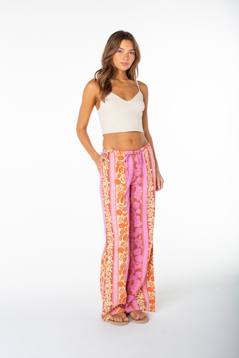 A woman stands against a white background in a cream crop top and Roxy’s Iyla Pant 2—wide-leg, high-waisted, with pink, orange, and cream floral stripes. Her hand is in her pocket, channeling effortless coastal style with chic sandals.