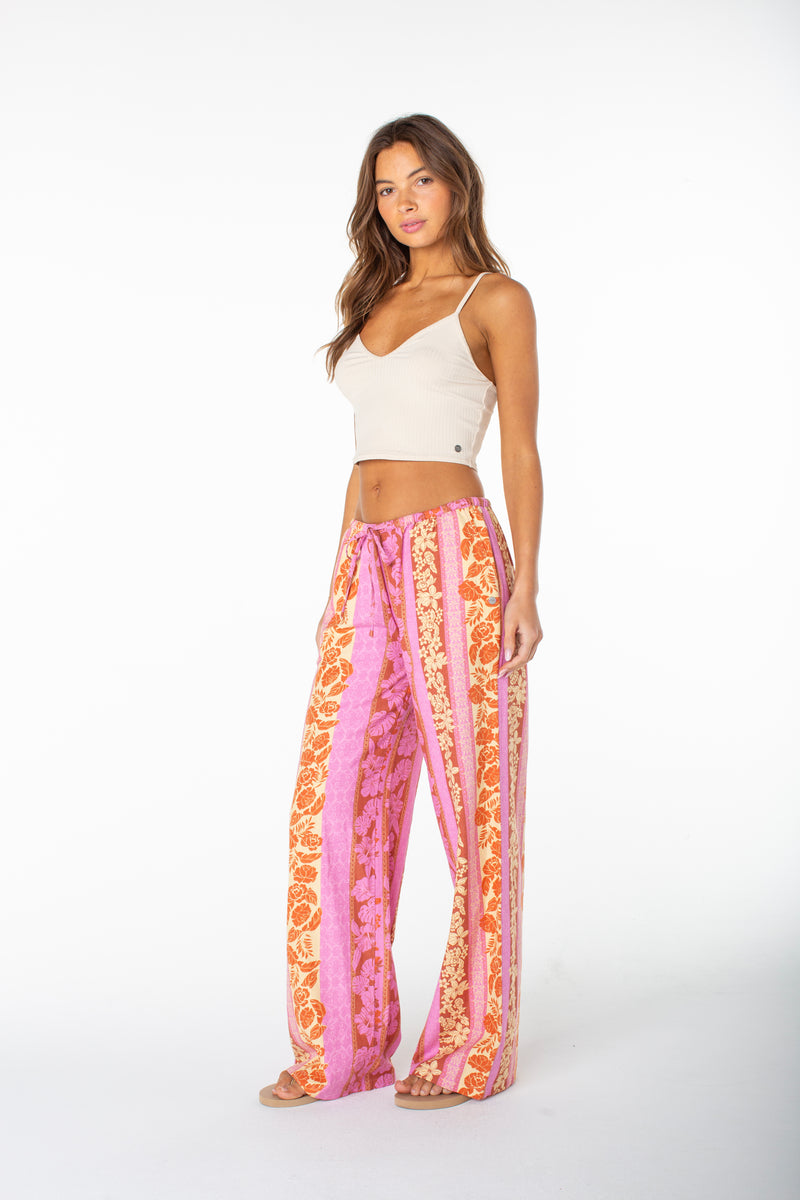 A woman stands against a white background wearing the Roxy Iyla Pant 2 in pink and orange patterns with a tie waist and a beige crop top, barefoot, exuding effortless coastal style as she looks confidently at the camera.