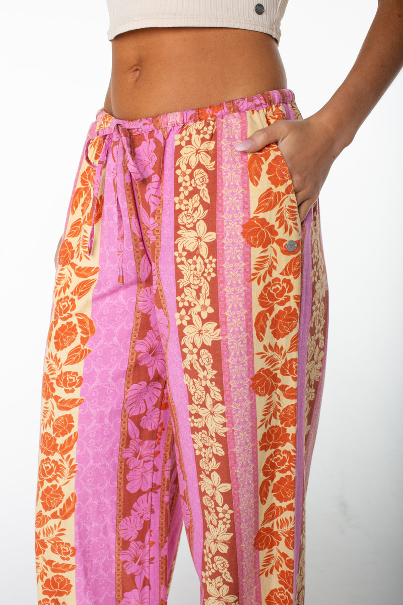 A person styles the Roxy Iyla Pant 2, featuring pink, orange, and yellow floral vertical stripes, a drawstring waist, and relaxed fit for coastal vibes; paired with a beige ribbed crop top and a hand in the pocket.