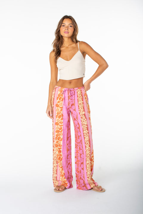 A woman stands in a white studio wearing Roxy's Iyla Pant 2 in Fuchsia Pink Lanai Stripe with a cream crop top, sandals, and her right hand on her hip, embodying relaxed coastal style. She has long hair and a neutral expression.