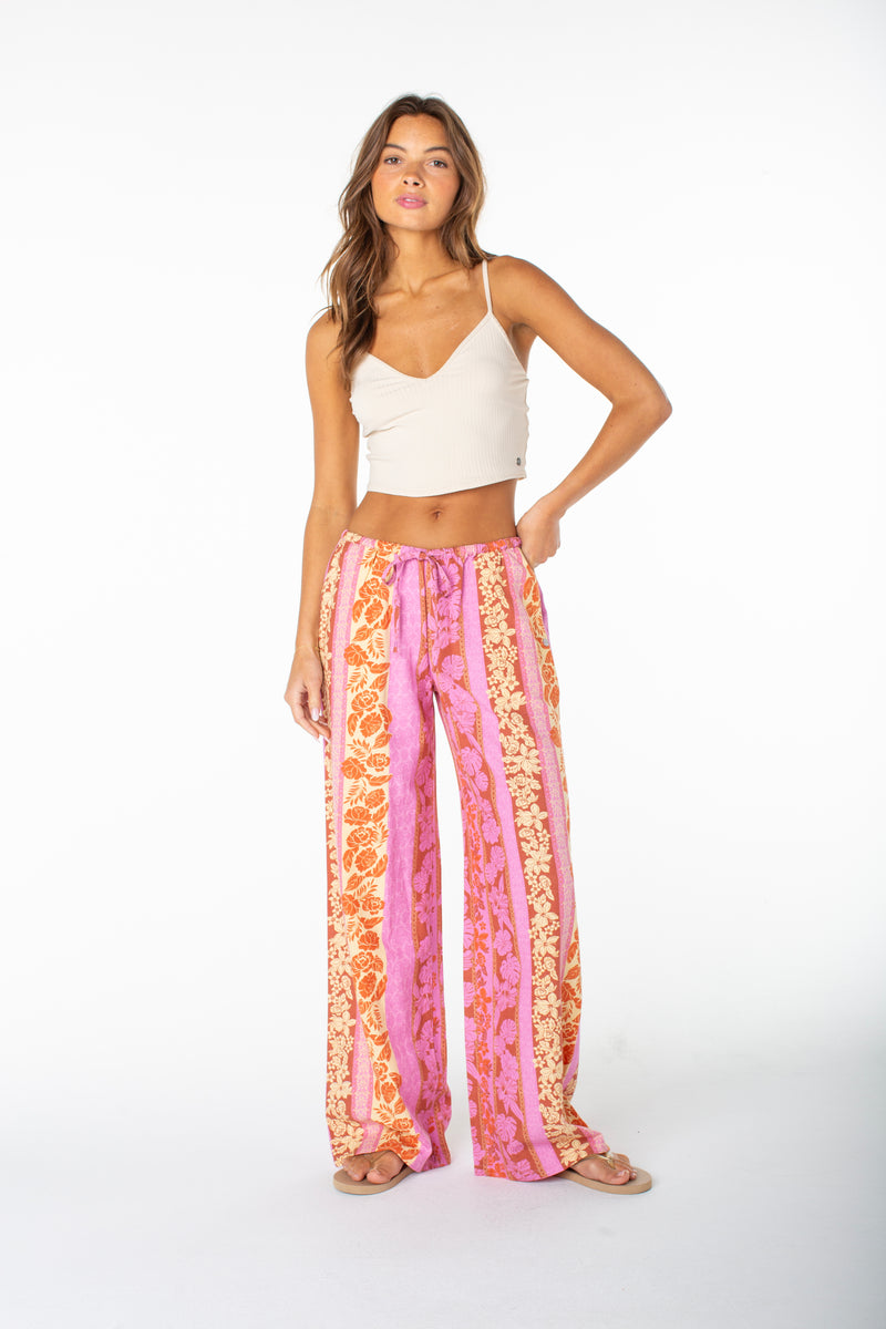 A woman stands in a white studio wearing Roxy's Iyla Pant 2 in Fuchsia Pink Lanai Stripe with a cream crop top, sandals, and her right hand on her hip, embodying relaxed coastal style. She has long hair and a neutral expression.