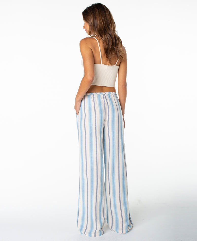 A woman stands facing away against a plain white background, wearing a sleeveless white crop top paired with the Roxy Iyla Pant 2—a relaxed-fit, high-waisted pant featuring blue and white vertical stripes—capturing an effortlessly coastal style.