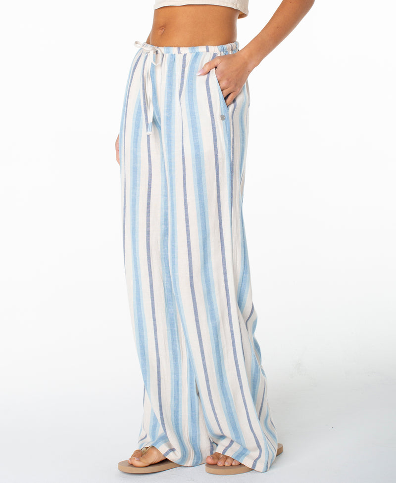 A woman models the Roxy Iyla Pant 2, a relaxed-fit, high-waisted pant featuring a wide-leg silhouette, vertical blue and white stripes, drawstring waist, and convenient pockets. Only her midsection and legs are visible against a plain white background, evoking an effortless coastal style.