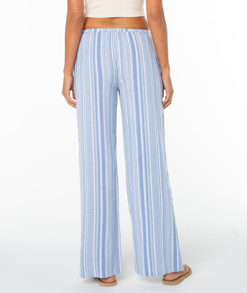 A person stands facing away against a white background, wearing the Roxy Iyla Pant 2 in light blue and white vertical stripes with a relaxed fit, styled with a white crop top and sandals for an effortless coastal look.