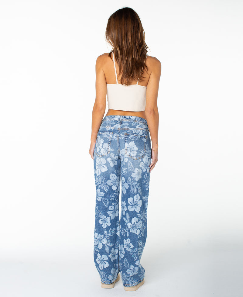 A woman with long brown hair, seen from the back and standing against a plain white background, wears a white crop top paired with high-waisted, relaxed-fit Haven Flora Pant by Roxy featuring a bold floral print.