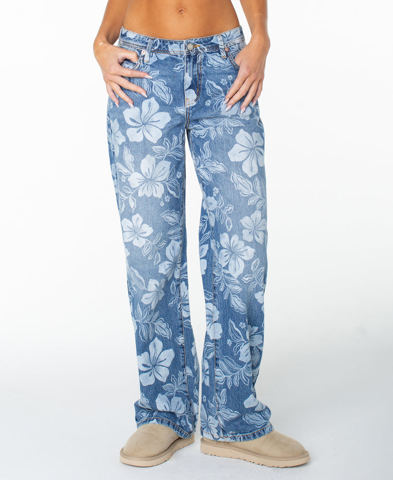A person wearing the Roxy Haven Flora Pant in blue, featuring a relaxed, oversized fit with a bold white hibiscus flower print, paired with tan slip-on shoes and a bare midriff. The background is plain white, and the image is cropped at the upper chest.