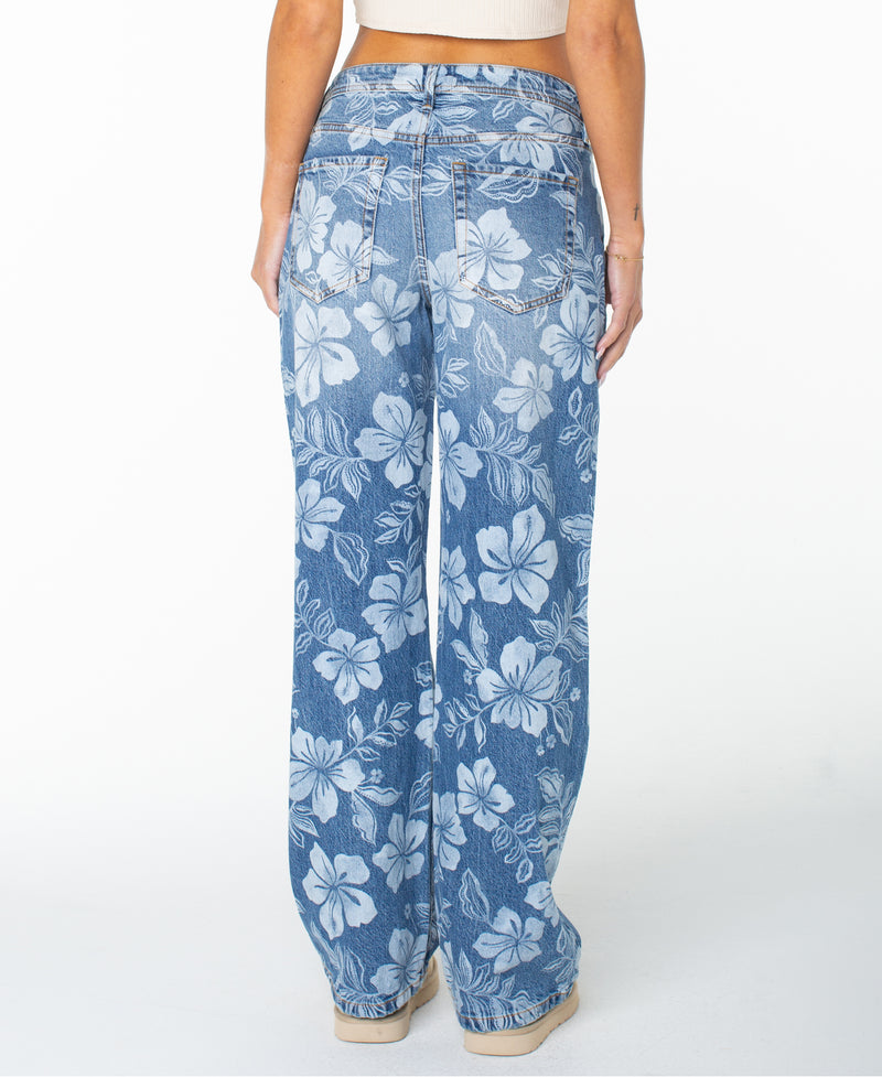 Model showcases the Roxy Haven Flora Pant, styled in a relaxed oversized fit with a wide-leg silhouette and bold white hibiscus floral print. Seen from the back, these pants feature two back pockets and tan stitching, effortlessly paired with beige shoes and a light crop top.