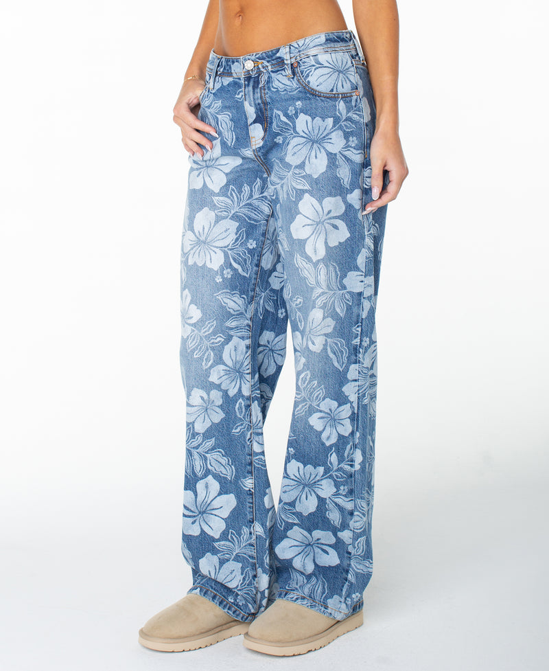 A person stands against a plain white background, wearing the Roxy Haven Flora Pant—high-waisted, wide-leg jeans featuring bold floral prints—paired with a bare midriff and beige slip-on shoes.