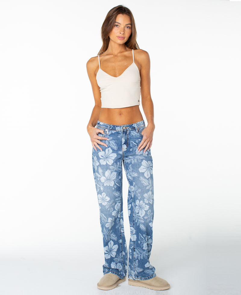 A woman stands on a plain white background, hands in her pockets, wearing a white crop top, the Roxy Haven Flora Pant in Flora Fade Print Denim Wash, and beige shoes.