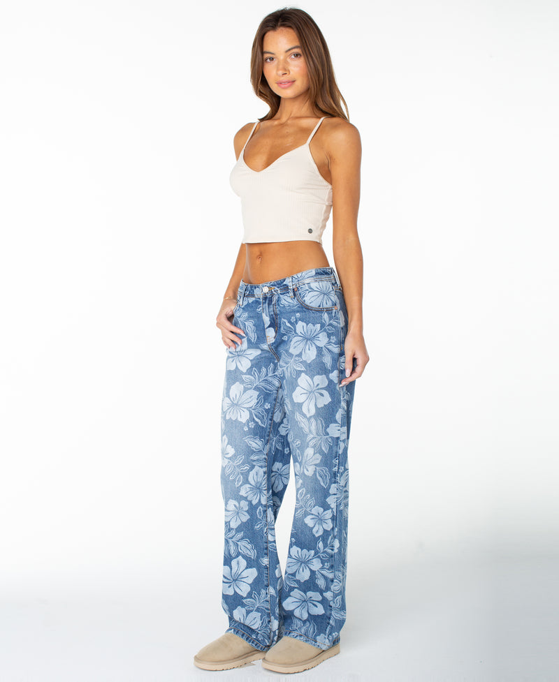 A woman stands against a white background wearing a beige crop top, the Roxy Haven Flora Pant—blue relaxed oversized jeans featuring a bold floral pattern—and beige shoes. She has long brown hair and is looking at the camera with a slight smile.