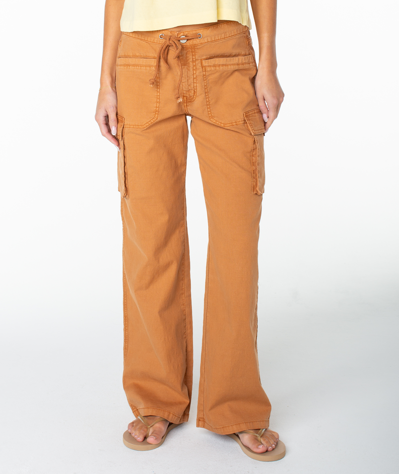 A person is shown from the shoulders down, wearing Roxy’s A La Cargo Pant in a low-waisted orange wash style with large pockets and a drawstring waist, paired with flip-flops, standing against a plain white background.
