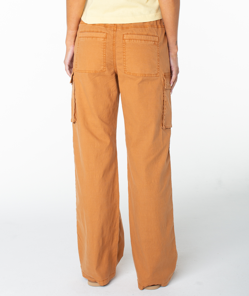 A person is seen from the back, standing against a plain white background, wearing the Roxy A La Cargo Pant—a pair of wide-leg, low-waisted cargo pants in a light brown and orange wash with large back and side pockets.