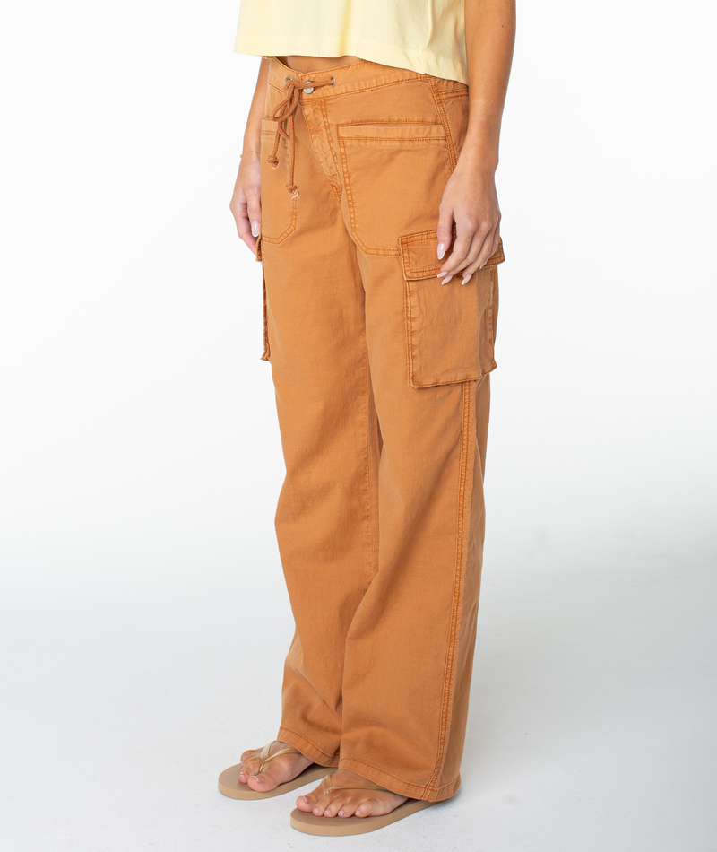 A person wearing Roxy’s A La Cargo Pant in Bran Solid, featuring side pockets and a drawstring waist, along with a light yellow top and beige flip-flops, stands against a plain white background.
