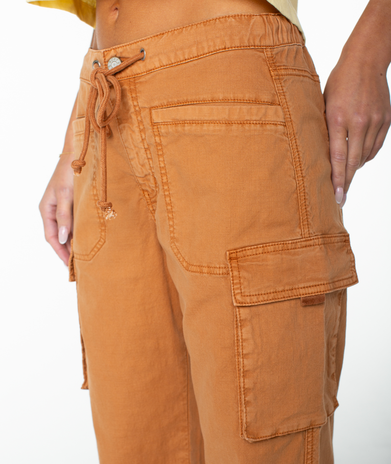 A person is shown from the lower torso down, modeling the Roxy A La Cargo Pant—low-waisted in an orange wash, featuring large front and side pockets, a drawstring waist, and visible stitching details—all set against a white background.