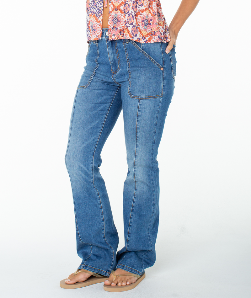 A person wearing Roxy's Trailhead Pant in blue bootcut, paired with a colorful patterned top and beige sandals, stands against a white background. Only the lower half is visible.