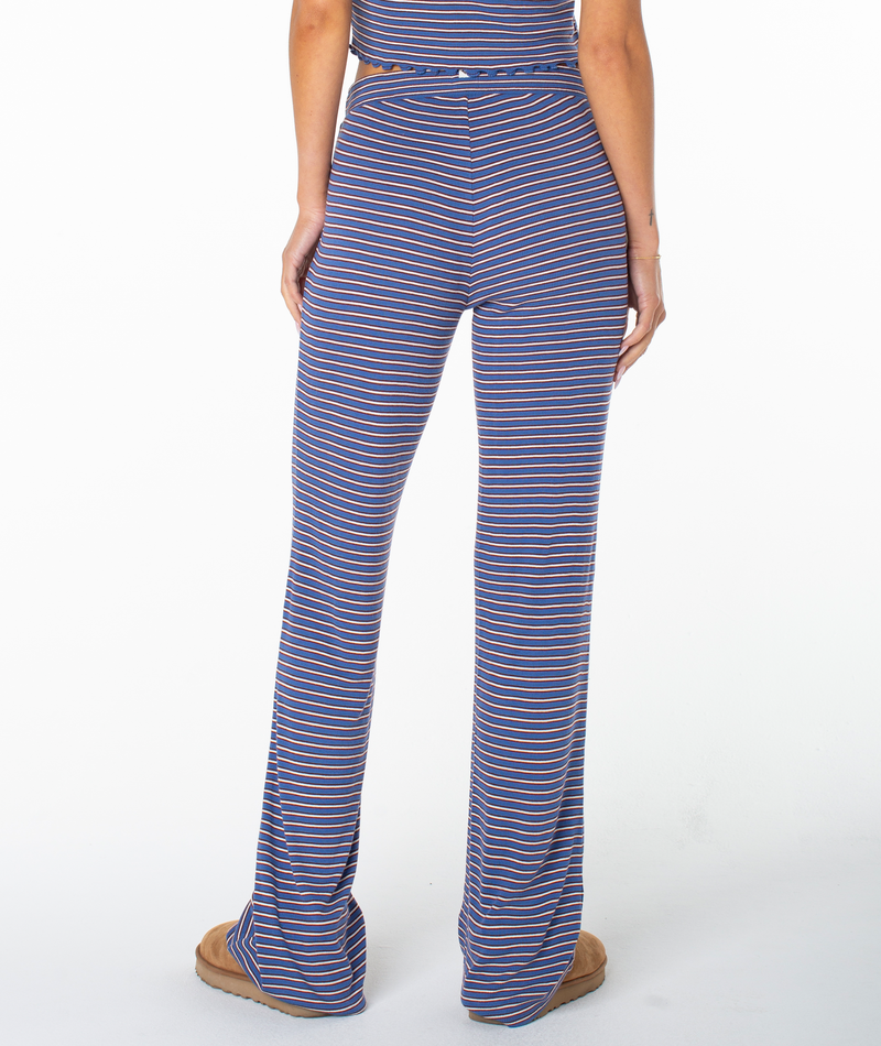 A person stands facing away, wearing the Roxy Summer Fling Rib Pant 2 in bold blue and white stripes paired with tan slippers. Their arms rest at their sides against a plain white background, capturing a hint of coastal flair.