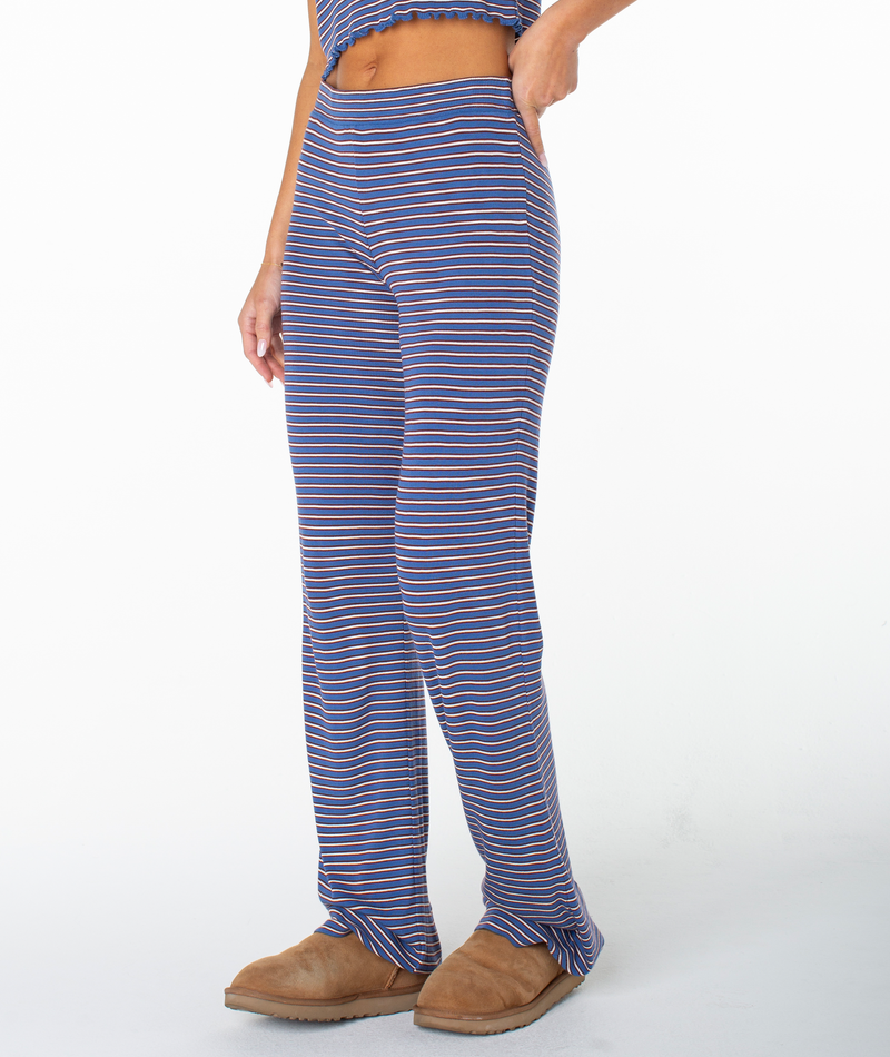 A person stands against a plain white background, exuding effortless coastal flair in the Roxy Summer Fling Rib Pant 2 featuring bold blue and white stripes, paired with a matching cropped top and brown slippers.