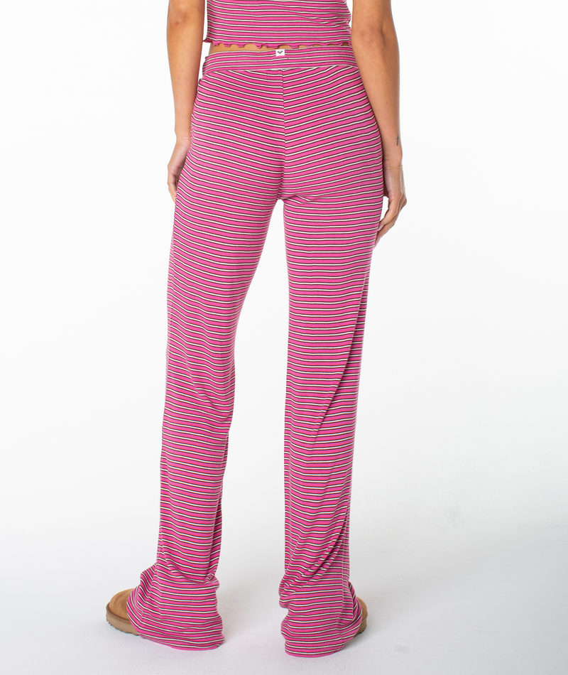 A person stands facing away from the camera, dressed in the Roxy Summer Fling Rib Pant 2. The pants feature bold pink and purple stripes, are long and loose-fitting, covering the shoes and adding a touch of coastal flair against the plain white background.