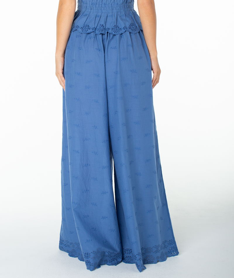A person wearing the Roxy Cambria Pant—a pair of high-waisted, wide-leg blue flowy pants with embroidered details at the hem—is photographed from the back against a white background. The relaxed tailoring of the Cambria Pant adds to its effortless appeal.