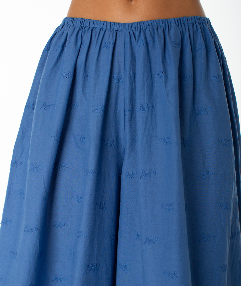 A close-up of a person wearing the Roxy Cambria Pant highlights its relaxed tailoring, featuring a blue, high-waisted design with subtle embroidered patterns. The upper body is not visible; the focus is on the textured fabric and gathered waistband.