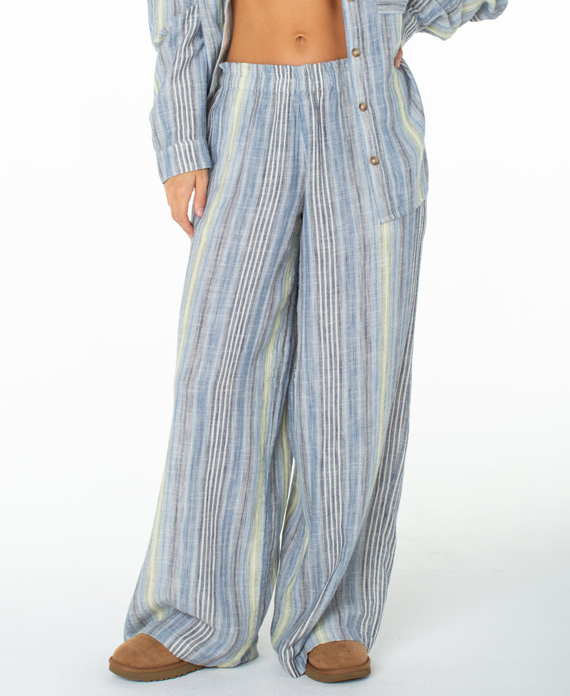 A person is seen from the midsection down wearing Roxy’s loose, striped blue and white Offshore Pant paired with a matching shirt. With one hand on their hip and brown slippers against a white background, this look captures easy vacation style.