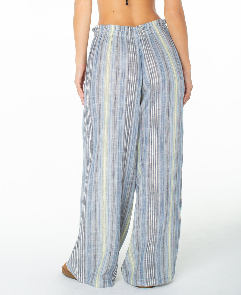 A person is shown from the back wearing the Roxy Offshore Pant—a pair of loose, wide-leg cotton pants in blue, gray, and yellow stripes. These vacation-ready pants feature an elastic waistband and a relaxed fit. The background is plain white.