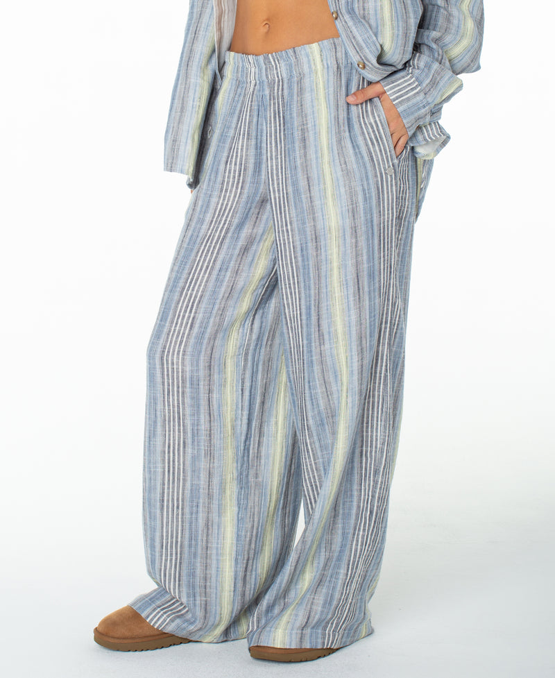 A person is seen wearing the Roxy Offshore Pant—loose, wide-leg pants featuring vertical blue, white, and yellow stripes in soft cotton for a laid-back, vacation-ready vibe—paired with a matching button-up shirt and brown shoes. The upper body is mostly out of frame.