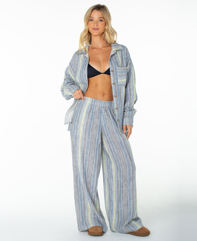 A woman stands smiling in vacation-ready Roxy Offshore Pants in Mood Indigo Coastline Stripe with a button-up shirt, black bra, and brown slippers against a white background.