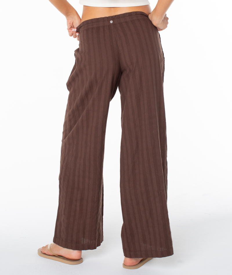 A person in loose, brown, striped cotton Offshore Pant by Roxy stands barefoot with one hand in their pocket, facing away from the camera against a white background—ideal for vacation.