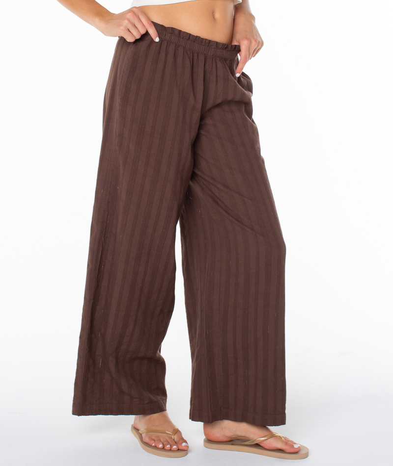 A person models the Roxy Offshore Pant in Chestnut Solid with beige flip-flops, standing against a white background and highlighting a relaxed, vacation-ready look.