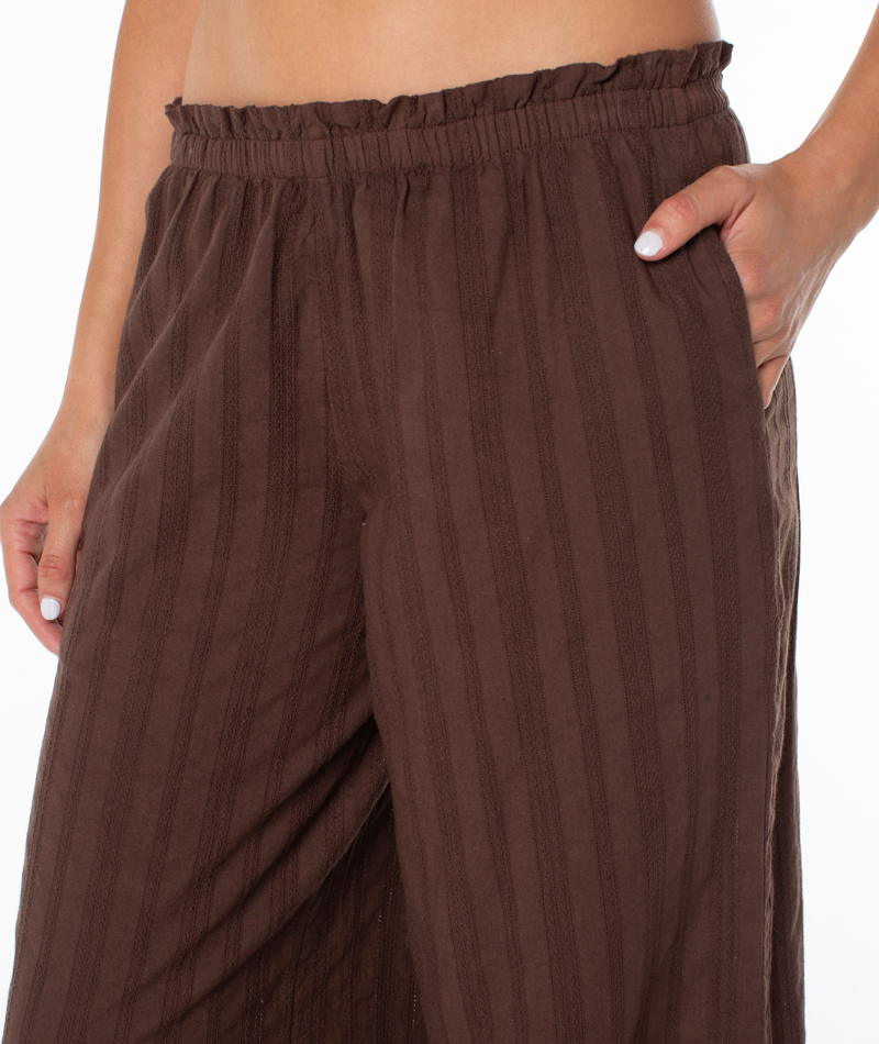 A person wears the vacation-ready Roxy Offshore Pant, featuring a high-rise stretchy ruffled waistband, side pocket, and wide-leg cotton fit—shown from waist to mid-thigh.