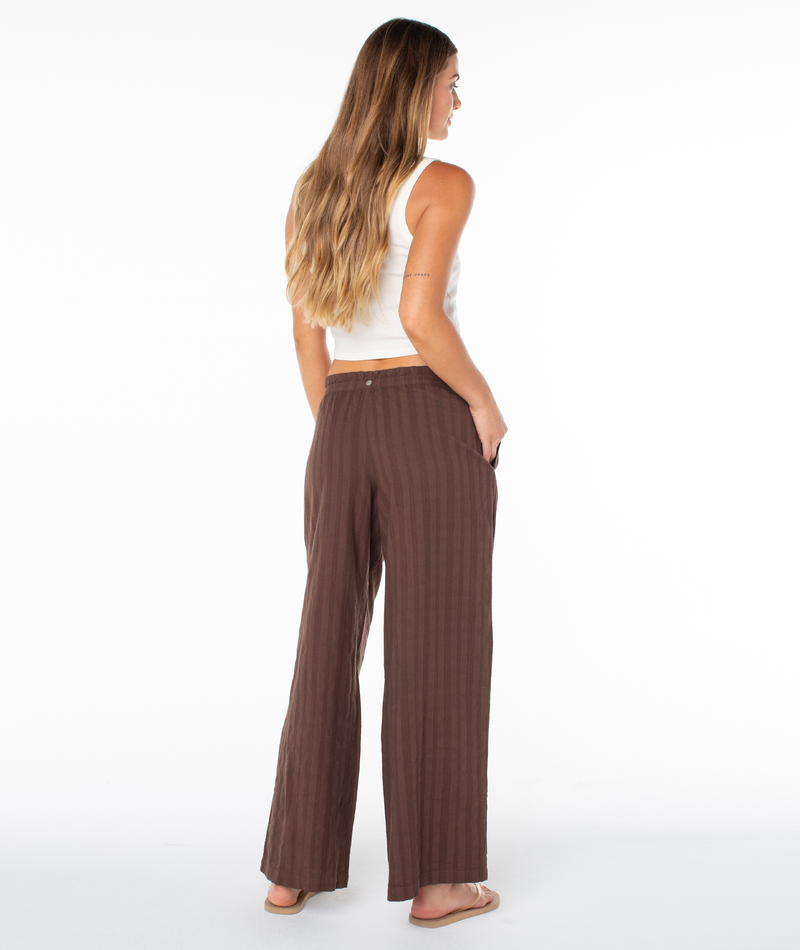 Woman with long hair in a white sleeveless top, Roxy Offshore Pant in brown, and sandals stands with her back to the camera against a white backdrop.