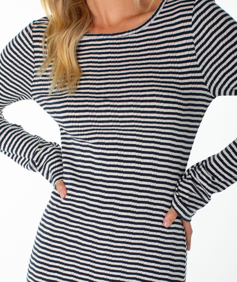 A person wearing the Roxy Hermosa Mini Dress, featuring long sleeves and black-and-white stripes in a cotton blend, stands with hands on hips. Only the upper body is visible, with light wavy hair cascading over one shoulder.