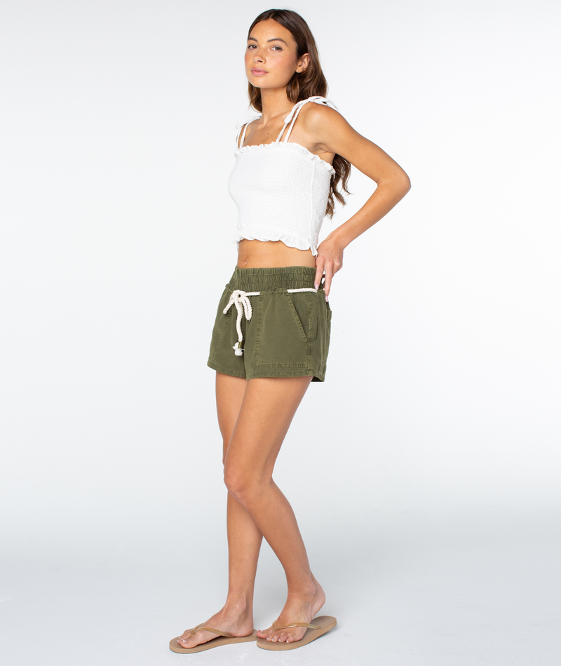 A woman poses in a white sleeveless crop top, Roxy Paddle On olive green linen blend drawstring shorts, and tan sandals against a plain white background.