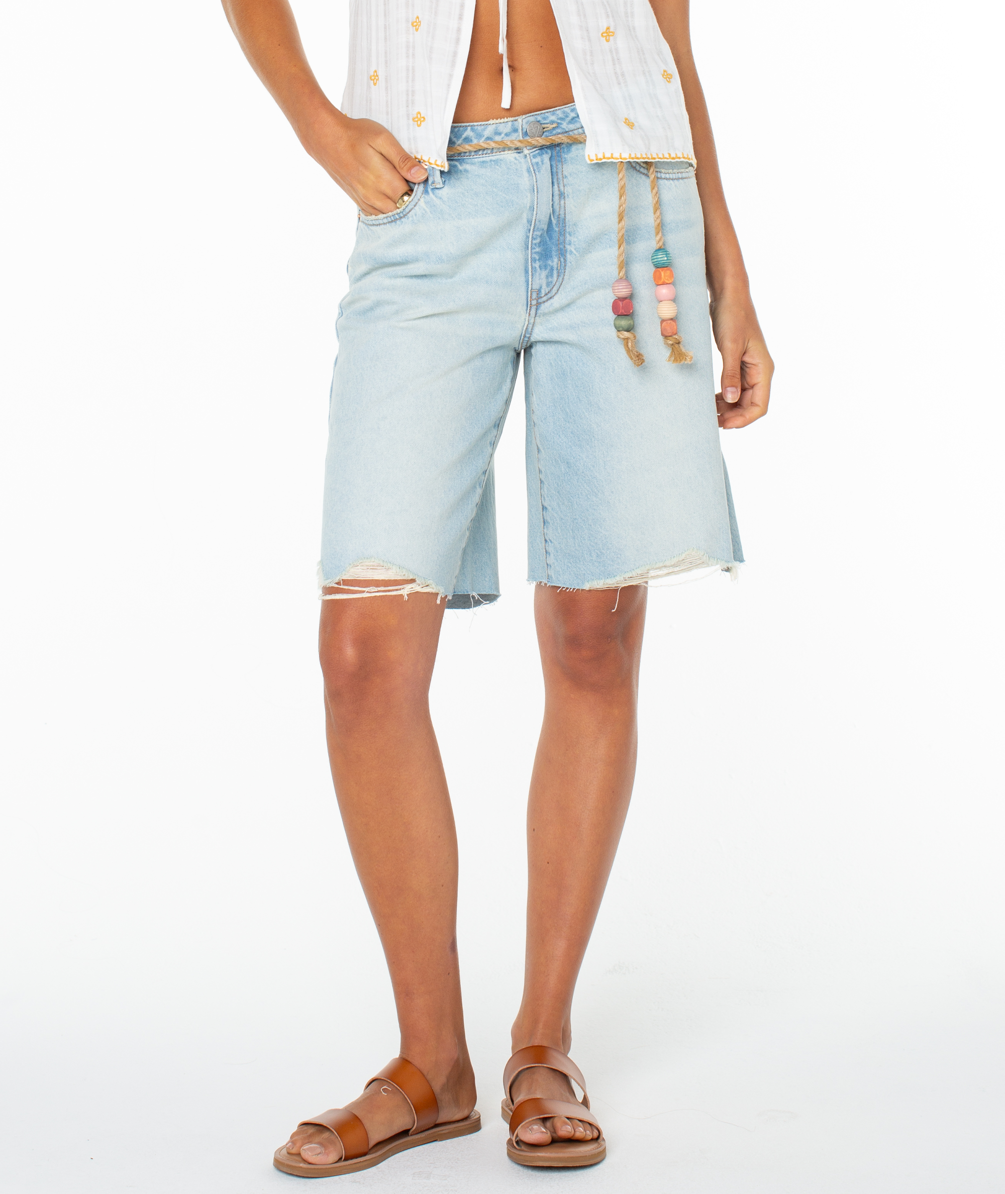 A person wearing Roxy's Message In A Bottle Short in light blue with frayed edges, paired with a white sleeveless top with rope tie and drawstring waist, plus brown sandals, stands against a white background.