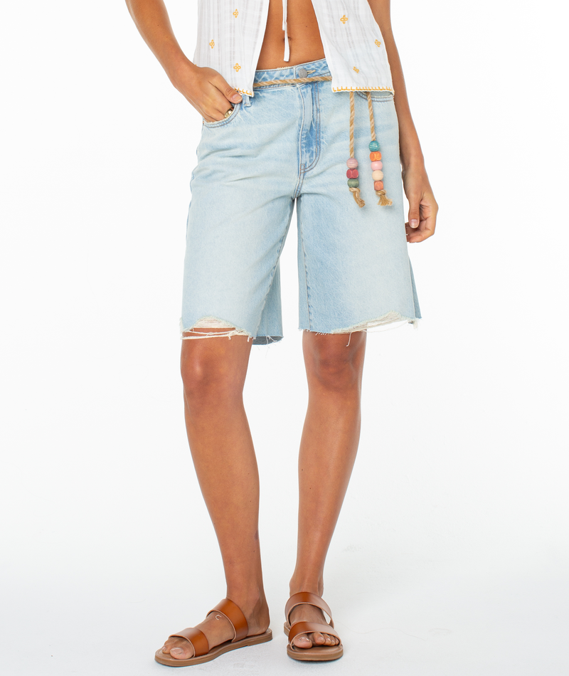 A person wearing Roxy's Message In A Bottle Short in light blue with frayed edges, paired with a white sleeveless top with rope tie and drawstring waist, plus brown sandals, stands against a white background.