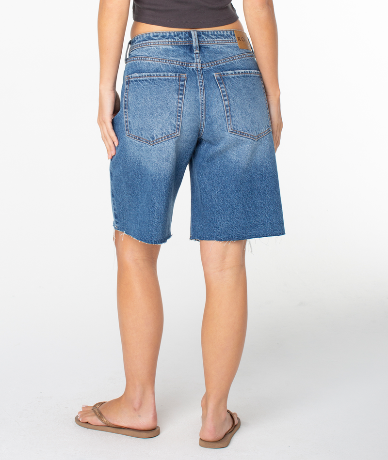 A person shown from the back wears the Roxy Lifesaver Short, a pair of blue midrise longline denim shorts with frayed hems, paired with a black tank top and brown flip-flops, set against a plain white background.