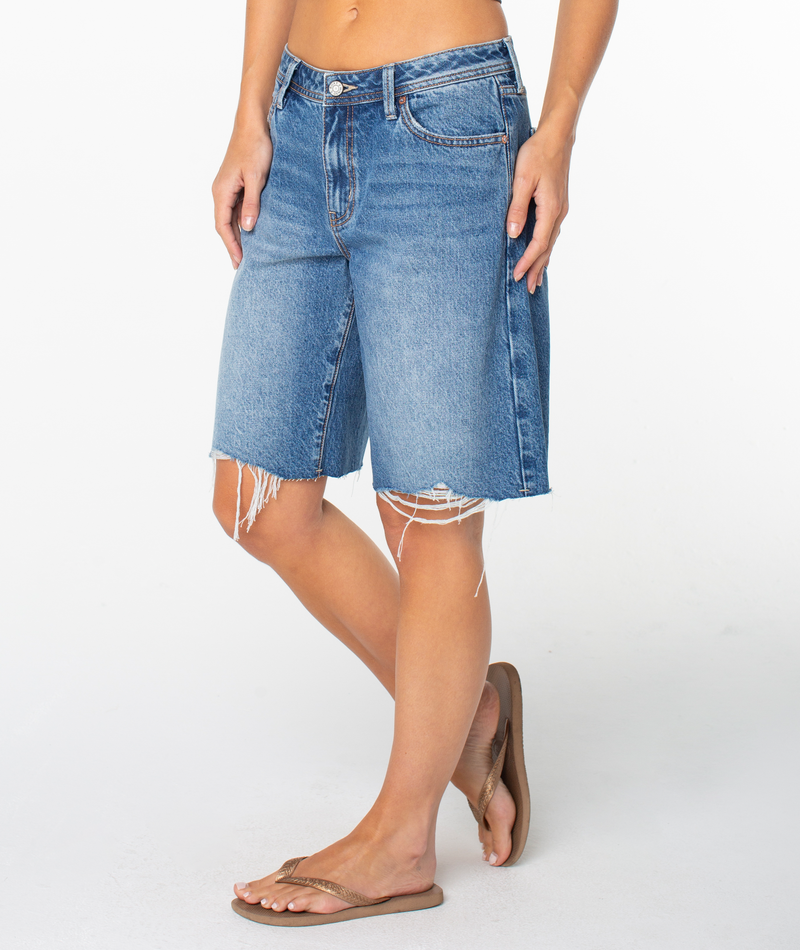 A person models the Roxy Lifesaver Short—knee-length, frayed-edge dark denim shorts with front and back pockets and a button-zipper closure—paired with brown flip-flops, against a plain white background.
