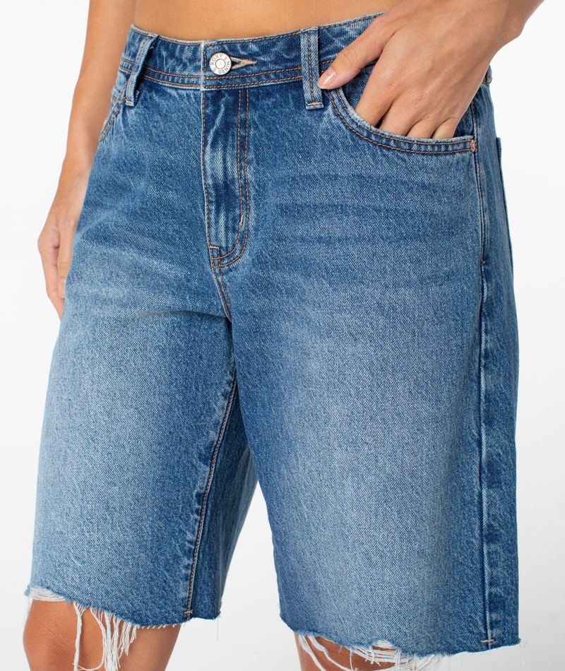 A person models the Roxy Lifesaver Short—midrise dark denim Bermuda shorts with frayed hems, one hand in a pocket—from waist to above the knees against a plain white background.