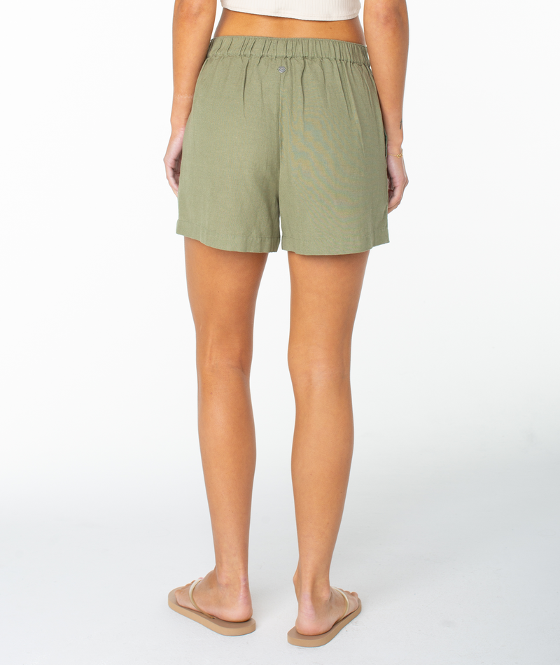 A person stands facing away against a plain white background, wearing the Roxy Mavericks Trouser Short in olive green, paired with a white crop top and beige sandals.