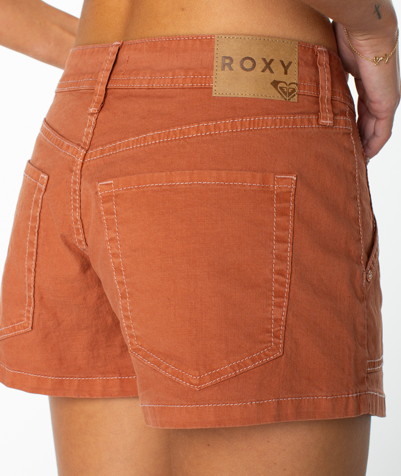 A close-up of a person wearing the Roxy Lost Treasure Short—relaxed, rust-colored denim shorts featuring visible stitching, carpenter pockets for added style, and a brown ROXY patch above the back pocket. Their hand rests gently by their side, highlighting the subtle coastal-inspired design.