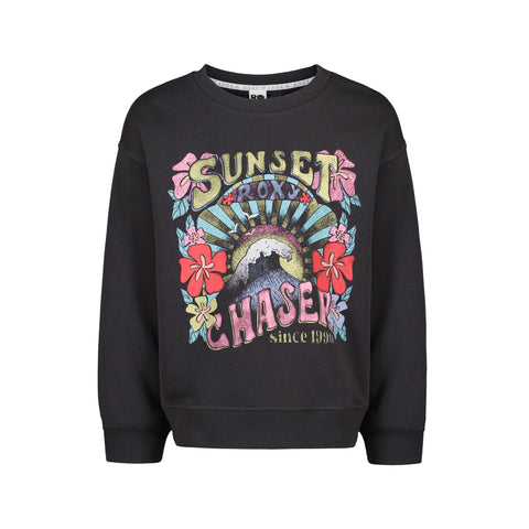 The Girls' Sunny Crew by Roxy is a black oversized sweatshirt with a vibrant front graphic—flowers, sunburst, wave, and “Sunset Chaser Roxy since 1990.” Made from soft cotton-poly fleece, pullover style. From the ROXY Sunny Crew collection.
