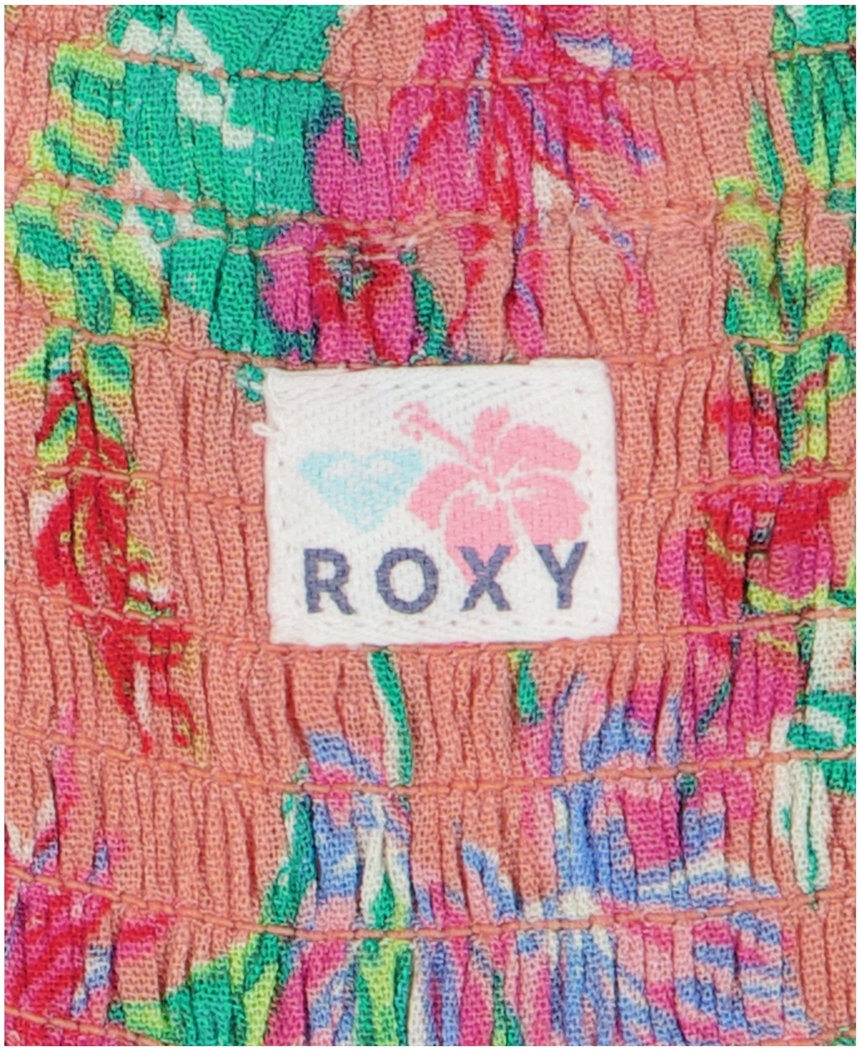 A close-up of peach fabric from the Girls' Coastal Breeze Jumpsuit by Roxy, featuring a green, red, and purple floral print. A white label reads ROXY beneath a pink hibiscus and blue diamond graphic.