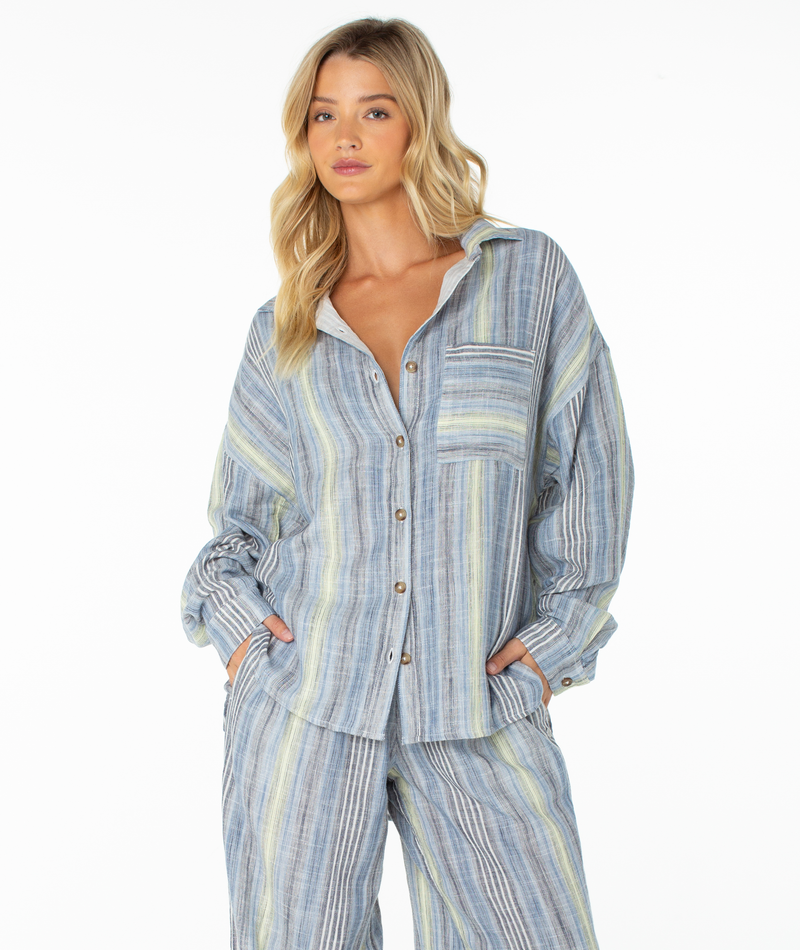A person wearing the Roxy Lets Get Away Top—a loose-fitting, blue and yellow striped shirt styled as a beach cover-up—along with matching pants, standing with hands in pockets against a white background.